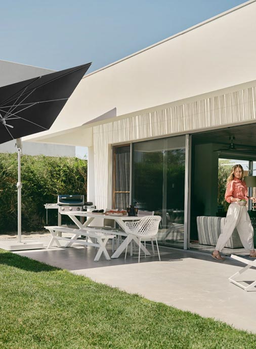 Eine leicht bekleidete Frau steht auf einer modernen Terrasse mit einem weißen Tisch und Stühlen unter einem großen schwarzen Sonnenschirm. Das Haus, das mit Burelbach Handwerk gebaut wurde, hat Glasschiebetüren, die sich zu einer sonnigen Grünfläche in Luxemburg und Deutschland öffnen.
