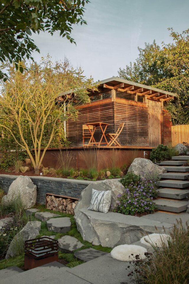 Ein modernes hölzernes Gartenhaus mit einer kleinen Terrasse und stilvollen Gartenmöbeln erhebt sich über einem begrünten Garten mit Steinstufen, Felsbrocken, Pflanzen und einem dekorativen Outdoor-Kissen, das ein durchdachtes Gardendesign präsentiert.