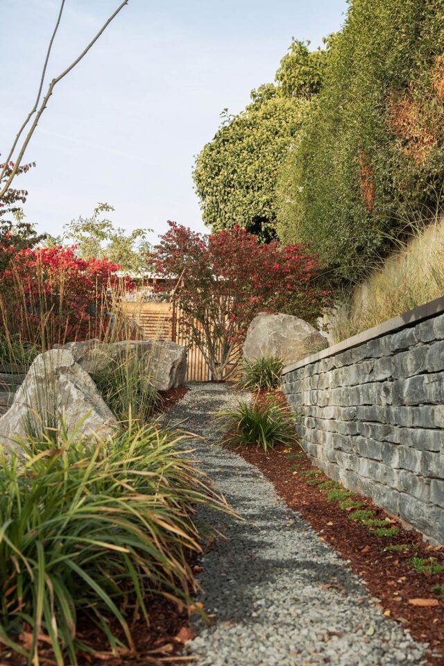 Ein von Ziergräsern, großen Steinen und Gartenmöbeln gesäumter Kiesweg führt zu einem hölzernen Tor, das von rot blühenden Sträuchern und hohen grünen Bäumen eingerahmt wird - ein ruhiger Rückzugsort, inspiriert von Burelbach in Deutschland und Luxemburg.