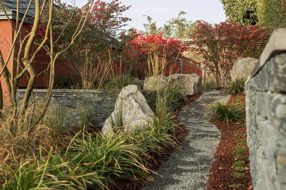 Ein geschotterter Gartenweg schlängelt sich an großen Steinen, Ziergräsern und blühenden Sträuchern vorbei und zeigt fachmännisches, von Deutschland und Luxemburg inspiriertes Gartendesign. Steinmauern säumen den Weg, und auf der linken Seite ist ein rotes Gebäude teilweise sichtbar.