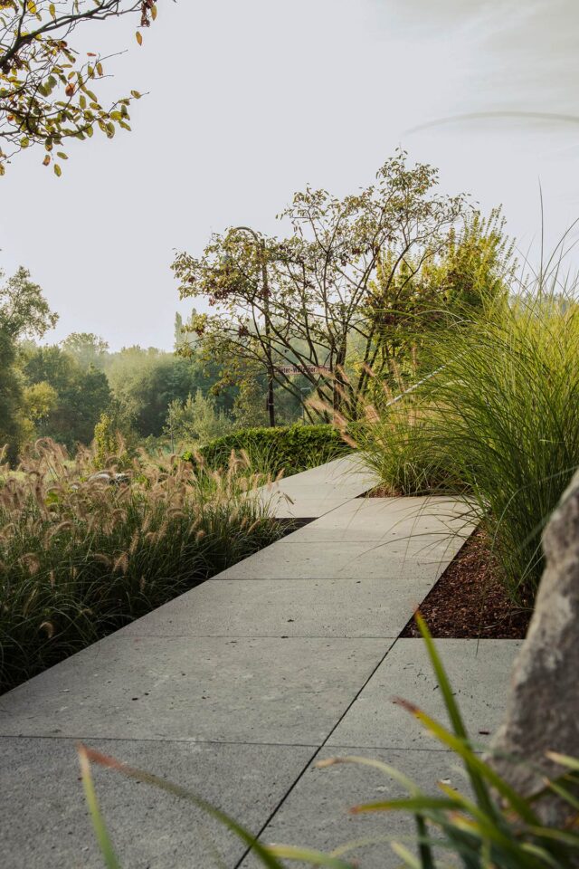 Ein betonierter Weg schlängelt sich durch hohe Gräser und Grünanlagen und zeigt fachmännisches Gardendesign mit üppigen Bäumen und Sträuchern im Hintergrund unter einem hellen, leicht bewölkten Himmel.