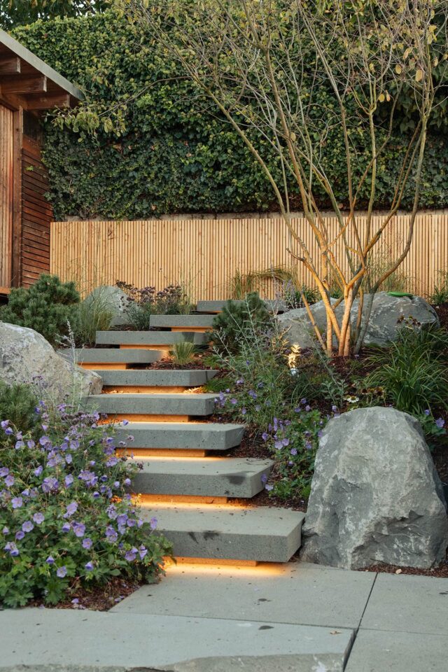 Betontreppen mit eingebauten Lichtern führen hinauf in einen von Burelbach Garten- und Landschaftsbau angelegten Garten, der von großen Steinen, lila Blumen und kleinen Bäumen gesäumt wird. Ein Bambuszaun und dichtes grünes Efeu vervollständigen diesen ruhigen Rückzugsort in Deutschland und Luxemburg.