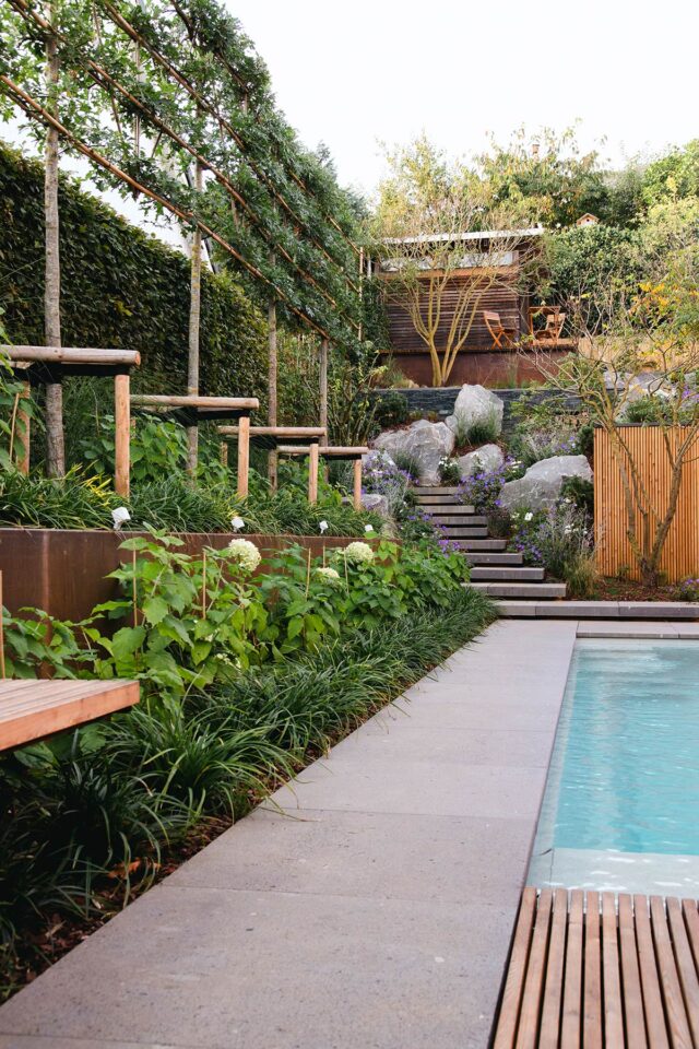 Moderner Garten von Handwerk Burelbach mit Swimmingpool, Steinweg, üppiger Bepflanzung, Holzbänken und terrassenförmigem Sitzplatz mit Stühlen und Tisch - alles fachmännisch in Deutschland und Luxemburg gefertigt.