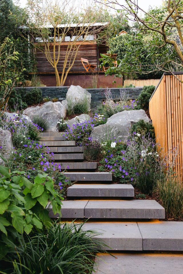 Steinstufen, gesäumt von üppigem Grün und violetten Blumen, führen hinauf zu einem hölzernen Gartenhaus mit einem orangefarbenen Stuhl in einer Garten- und Landschaftsbauanlage von Burelbach, umgeben von Bäumen und Felsen - ein friedlicher Rückzugsort in Deutschland und Luxemburg.