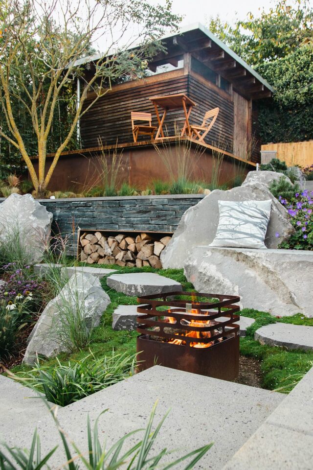 Ein moderner Garten von Burelbach Handwerk mit einem kleinen Holzschuppen, einem Tisch und Stühlen auf einer erhöhten Terrasse, gestapeltem Brennholz, einer Feuerstelle aus Metall, großen Felsbrocken, Steintreppen und üppigem Grün im gesamten angelegten Garten.