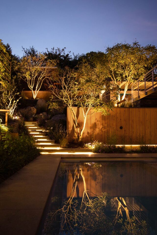 Ein wunderschön beleuchteter Garten in der Abenddämmerung mit einer Treppe, die durch Bäume und Pflanzen neben einem Pool hinaufführt, wobei der Weg und der Zaun mit warmen Lichtern angestrahlt werden - ein wahres Musterbeispiel für exquisites Handwerk in Burelbach.