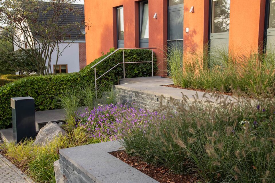 Vordereingang eines modernen Hauses mit orangefarbenen Wänden, grauen Türen, Steinstufen und Gartengestaltung von Burelbach. Ziergräser, violett blühende Pflanzen und ein Metallgeländer akzentuieren diesen stilvollen Look in Deutschland und Luxemburg.
