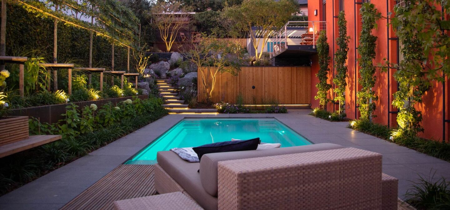 Ein moderner Garten von Burelbach Garten- und Landschaftsbau bietet einen kleinen beleuchteten Pool, Liegestühle, üppiges Grün und warme Landschaftsbeleuchtung - und setzt exquisites Handwerk für eine gemütliche und einladende Abendatmosphäre in Szene.