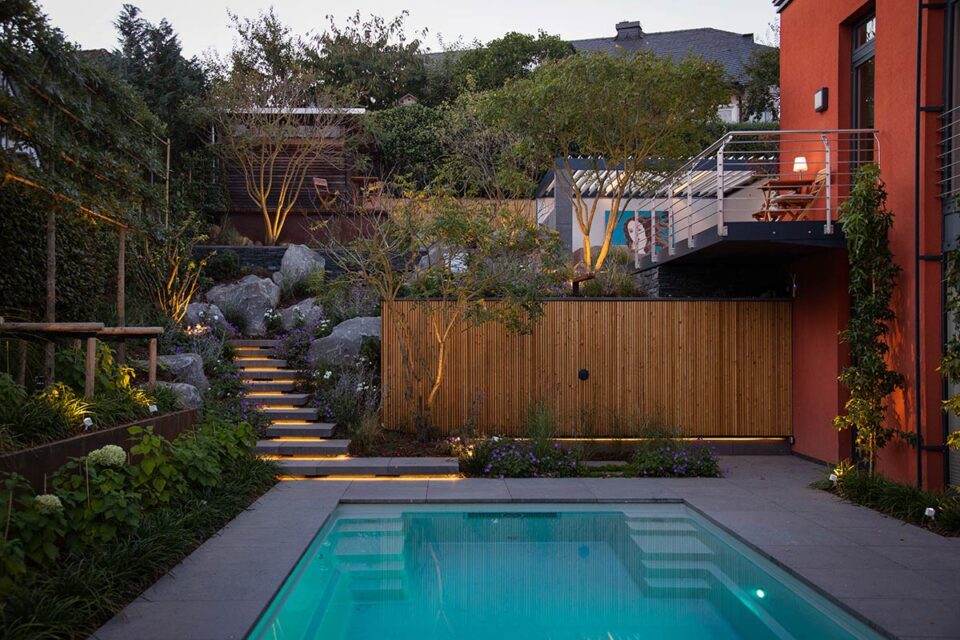 Moderner Hinterhof mit beleuchtetem Swimmingpool, terrassiertem Garten, stilvollen Gartenmöbeln von Burelbach, beleuchteten Stufen und einem Holzzaun; eine Person steht auf dem oberen Balkon des Orangenhauses. Perfekt für Abende in Luxemburg und Deutschland.