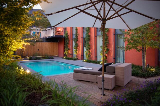 Moderner Hinterhof mit rechteckigem Pool, gepolsterten Gartenmöbeln auf einer Holzterrasse, Sonnenschirm, Pflanzen und einem orangefarbenen Gebäude mit hohen Fenstern im Hintergrund, abends warm beleuchtet - perfekt für Garten- und Landschaftsbau in Luxemburg und Deutschland.