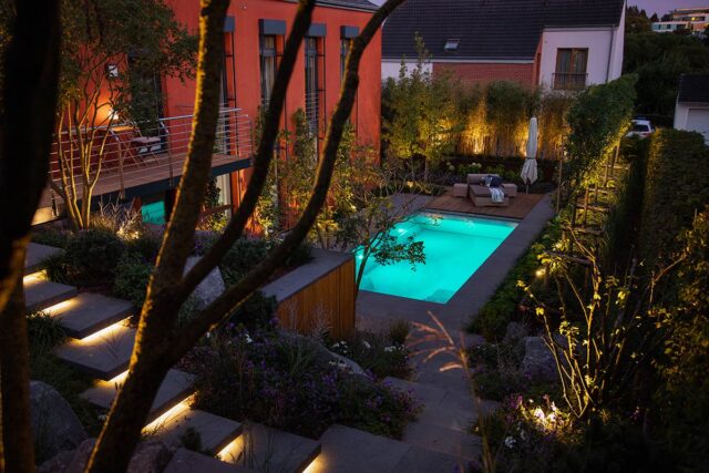 Ein moderner Garten in der Abenddämmerung zeigt fachmännisches Gardendesign mit einem leuchtenden rechteckigen Pool, stimmungsvoller Wegbeleuchtung, Bäumen, Sträuchern und stilvollen Gartenmöbeln in der gemütlichen Sitzecke, alles umgeben von modernen Häusern.