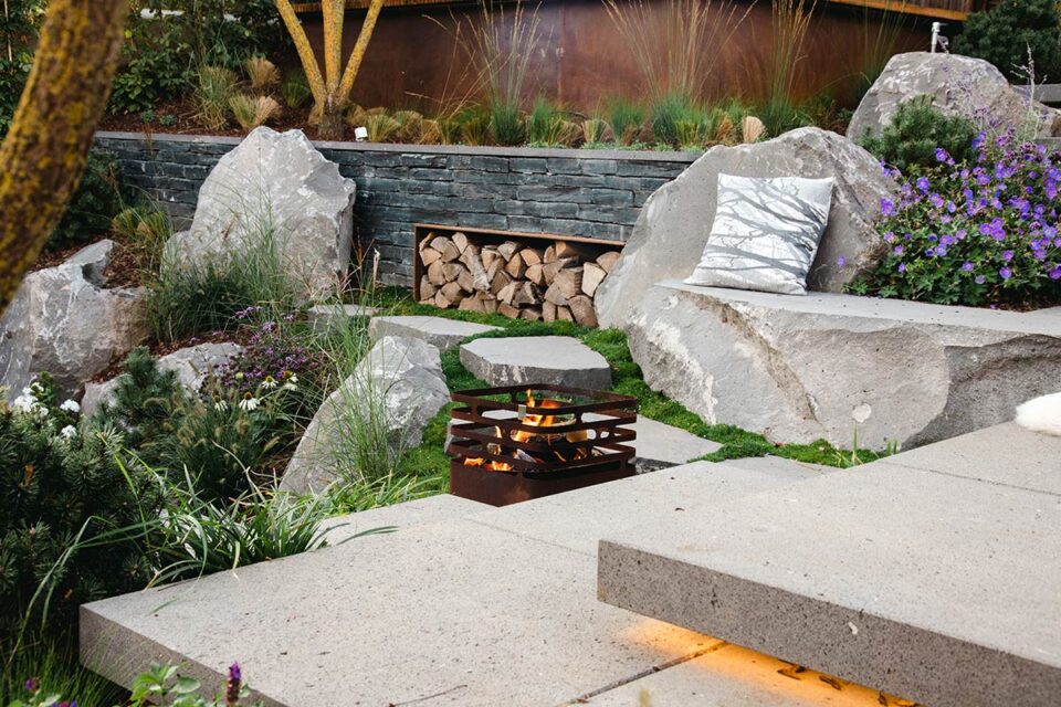 Eine moderne Gartengestaltung mit großen Steinen, üppigen Pflanzen, einem Stapel Brennholz, einem dekorativen Kissen und einer kleinen Metallfeuerstelle auf einer Steinterrasse mit Stufen und Akzentbeleuchtung.