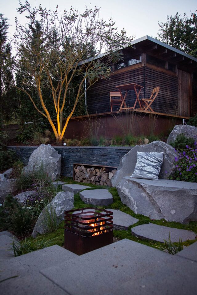 Ein gemütlicher Garten mit großen Steinen, einer kleinen Feuerstelle, einem Haufen Brennholz, handwerklich inspirierten Gartenmöbeln und einem Holzschuppen mit Tisch und Stühlen, alles umgeben von Pflanzen und Bäumen in der Abenddämmerung.