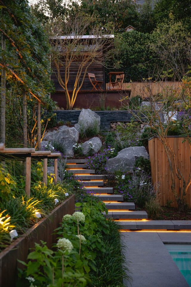 Eine Gartentreppe mit eingebauten Lichtern, die vom Fachmann für Garten- und Landschaftsbau angefertigt wurde, führt an üppigen Pflanzen und Steinen vorbei zu einer gemütlichen Holzterrasse mit zwei Stühlen, die in der Dämmerung sanft beleuchtet und von Bäumen und einem Zaun umgeben ist.