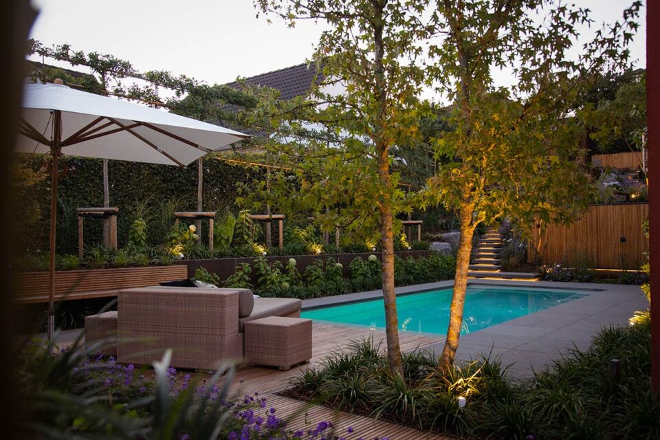 Ein moderner Garten mit fachmännischem Gartendesign mit rechteckigem Pool, Holzdeck, Sitzgelegenheiten im Freien, Sonnenschirm, üppigen Pflanzen und einem Baum; Stufen führen zu einem höher gelegenen Gartenbereich, der von handgefertigtem Garten- und Landschaftsbau eingerahmt wird.