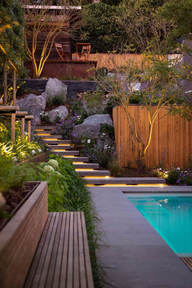 Ein moderner Garten mit handgefertigten Gartenmöbeln, beleuchteten Steinstufen, die zu einer Sitzecke führen, üppigem Grün, Felsen und einem türkis beleuchteten Pool. Die warme Beleuchtung schafft eine gemütliche Atmosphäre - perfekt für Häuser in Luxemburg und Deutschland.