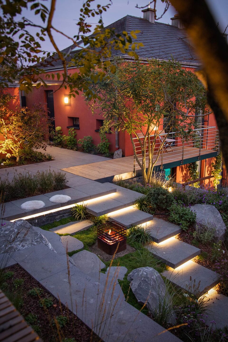 Ein modernes Gardendesign in der Abenddämmerung mit beleuchteten Steinstufen, die zu einer Terrasse führen. Eine kleine Feuerstelle befindet sich neben dem Weg, umgeben von Grün und Steinen. Warme Außenbeleuchtung beleuchtet das Haus und den Balkon, während stilvolle Gartenmöbel zum Entspannen einladen.