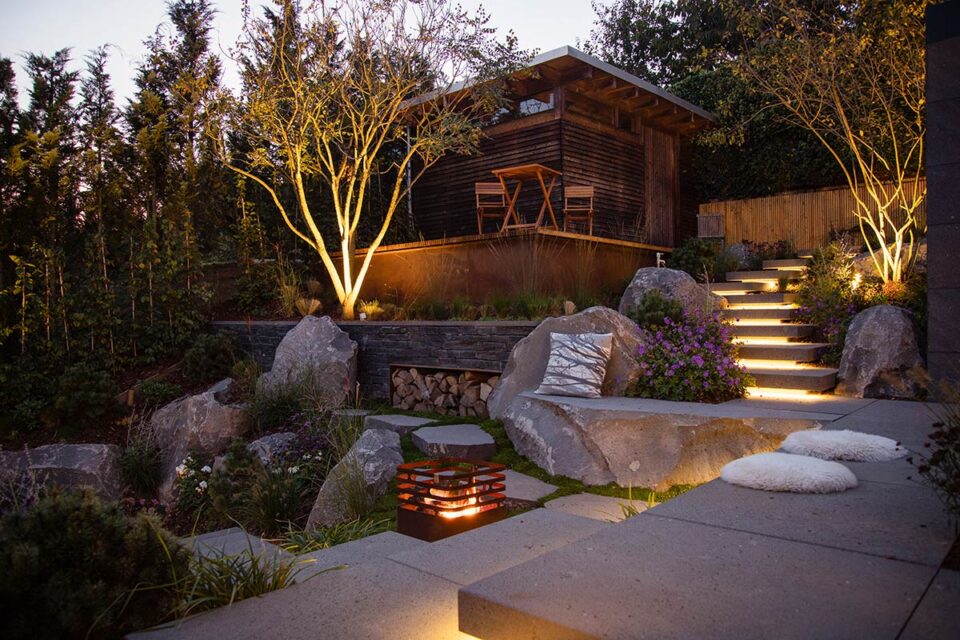 Ein in der Abenddämmerung angelegter Garten mit leuchtenden Lichtern, Steinstufen, großen Felsen und einem gemütlichen Sitzbereich - ein perfektes Beispiel für Garten- und Landschaftsbau. Kissen und eine beleuchtete moderne Feuerstelle sorgen für Wärme in diesem einladenden Außenbereich.