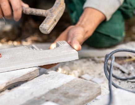 Eine Person übt das Handwerk aus, indem sie einen Hammer benutzt, um Fliesen oder Steine zu bearbeiten - sorgfältig umgeben von anderen Fliesen und einem Stromkabel -, was auf Bau- oder Renovierungsarbeiten in Luxemburg und Deutschland hindeutet, möglicherweise im Zusammenhang mit Gardendesign-Projekten.