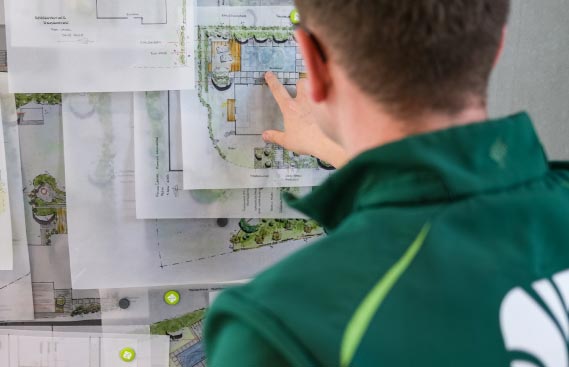 Eine Person in einer grünen Jacke zeigt auf Architekturpläne oder Blaupausen, die auf einem Tisch liegen, und prüft detaillierte Entwürfe und Diagramme für eine innovative Gartengestaltung.