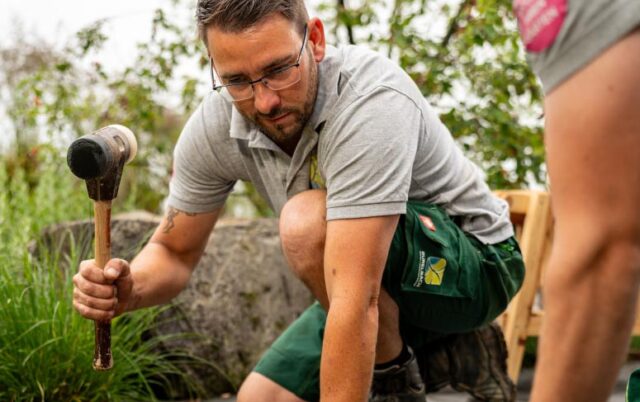 Ein Mann mit Brille kniet in Luxemburg im Freien, hält einen Hammer in der Hand und arbeitet an einem Outdoormöbel-Projekt.