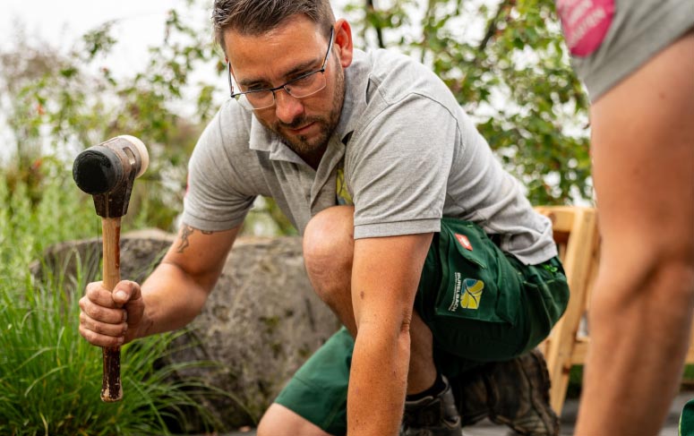 Ein Mann mit Brille und Arbeitskleidung kniet im Freien, hält einen Hammer in der Hand und arbeitet an einem handwerklichen Landschaftsbau-Projekt. Eine weitere Person steht in der Nähe. Im Hintergrund sind Grünflächen, Felsen und Elemente von Gardendesign zu sehen.