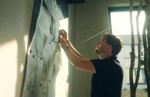 Ein Mann mit Brille und schwarzem Hemd heftet in einem sonnendurchfluteten Raum, in dem große Fenster und ein Ast im Hintergrund zu sehen sind, Papiere an eine Tafel. Er plant Garten- und Landschaftsbauprojekte für Burelbach in Luxemburg und Deutschland.