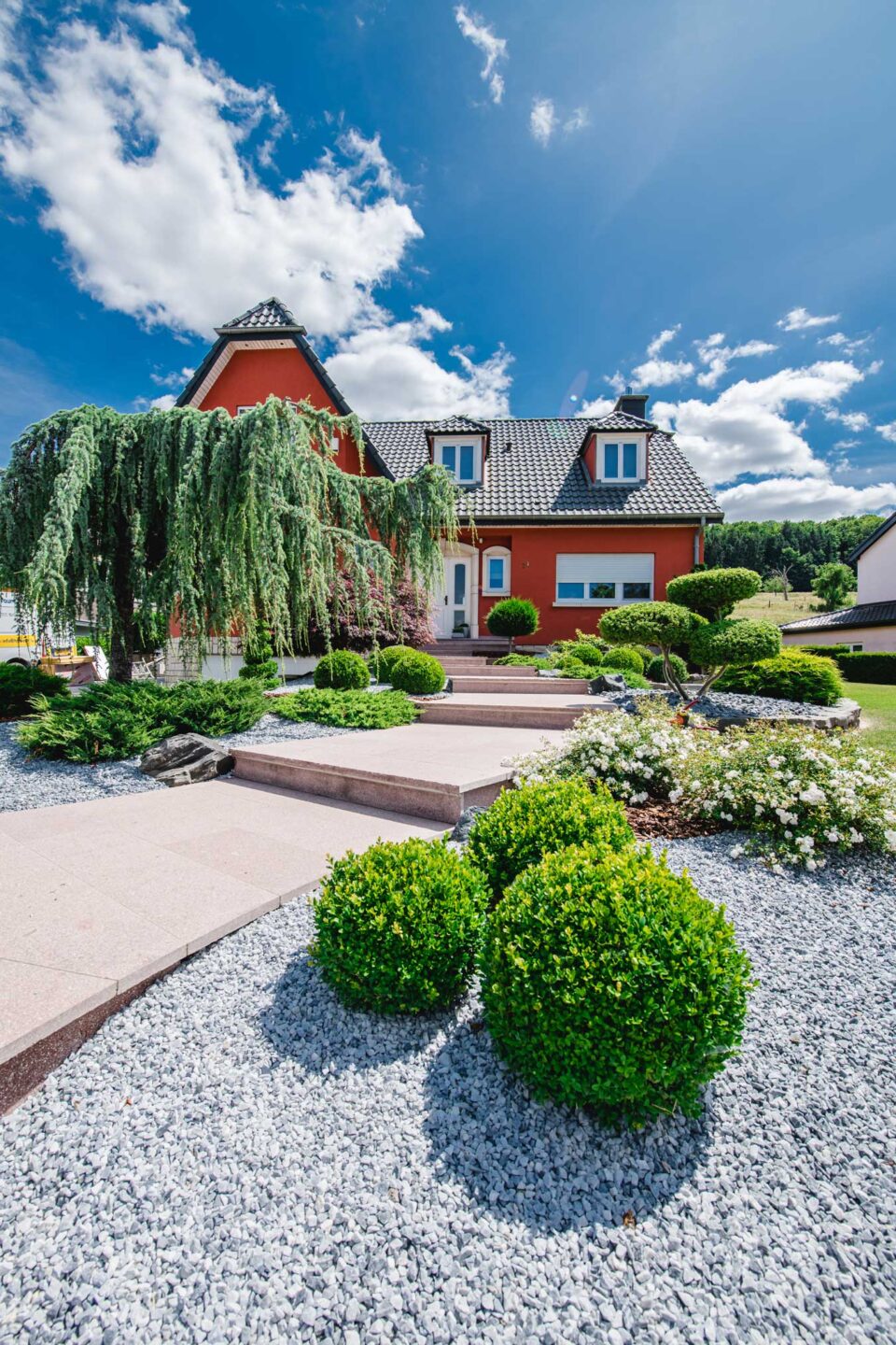Ein rotes Haus mit weißer Umrandung liegt hinter einem handwerklich hervorragend gestalteten Garten mit gestutzten Sträuchern, Kies, blühenden Pflanzen und einem Steinweg - ein Beispiel für Gardendesign aus Luxemburg und Deutschland unter strahlend blauem Himmel.