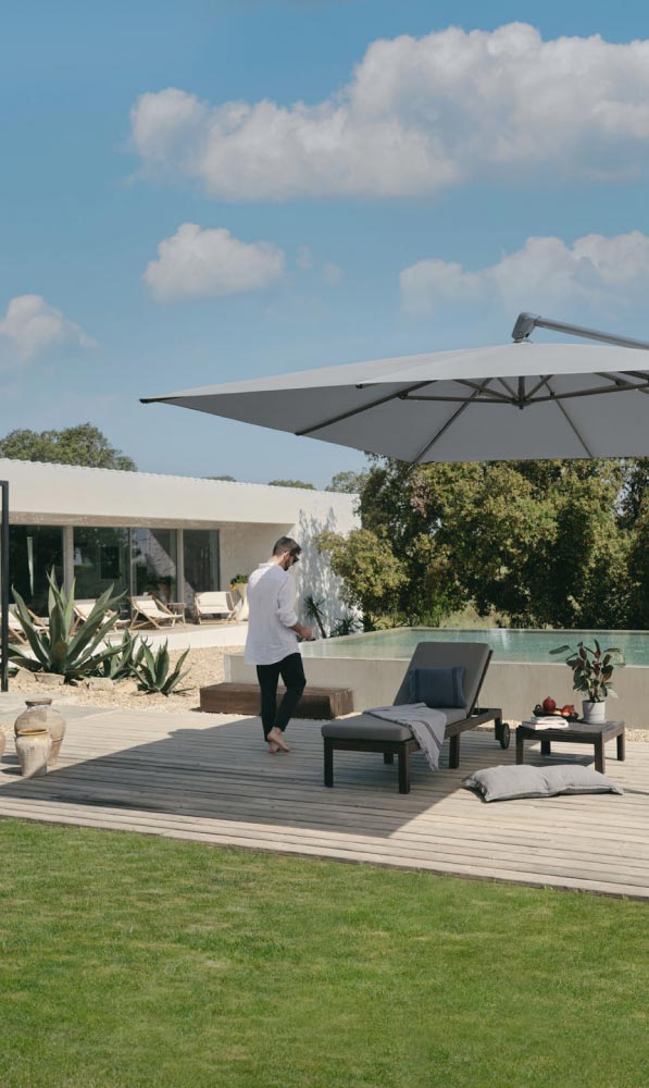 Eine Person läuft barfuß auf einer Holzterrasse neben einem Pool mit Liegestühlen, einem Tisch und einem großen Sonnenschirm - ein Beispiel für elegantes Gardendesign, inspiriert von moderner Architektur und Grün in Luxemburg und Deutschland, bei teilweise bewölktem Himmel.
