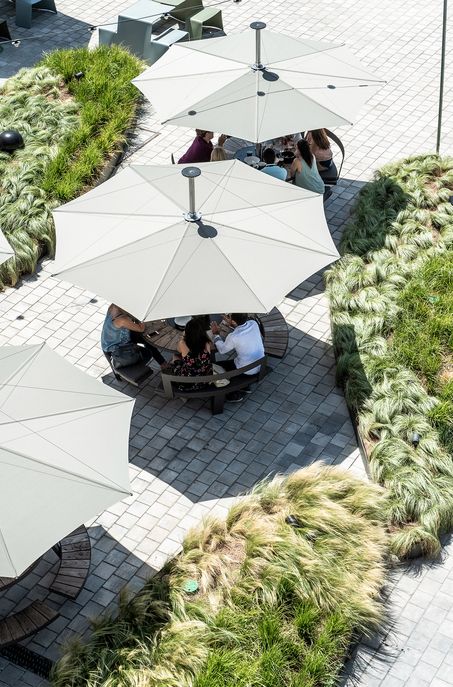 Die Menschen sitzen an Tischen unter großen weißen Sonnenschirmen auf einem Platz mit gepflasterten Wegen und begrünten Flächen, die ein elegantes, von Luxemburg und Deutschland inspiriertes Gardendesign zeigen, von oben betrachtet.