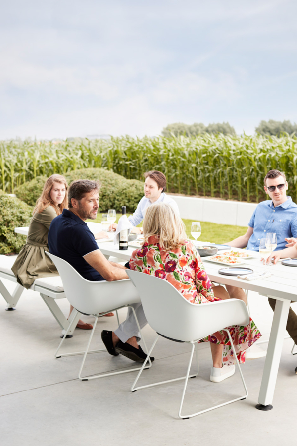 Sechs Personen sitzen um einen Esstisch im Freien mit Speisen und Getränken und unterhalten sich. In der Nähe eines grünen Maisfeldes und unter blauem Himmel genießen sie die von Gardendesign inspirierte Umgebung für ein entspanntes, zwangloses gemeinsames Essen.