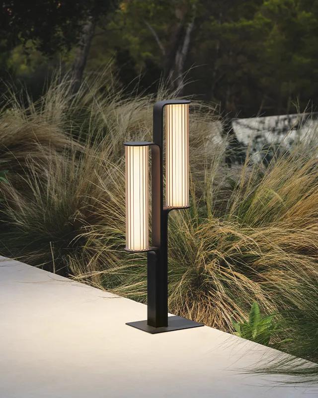 Eine moderne Außenlampe mit zwei vertikalen, rechteckigen Leuchten steht neben einem Betonweg, umgeben von hohem Gras und Grün - ein eleganter Touch von Gardendesign by Burelbach.