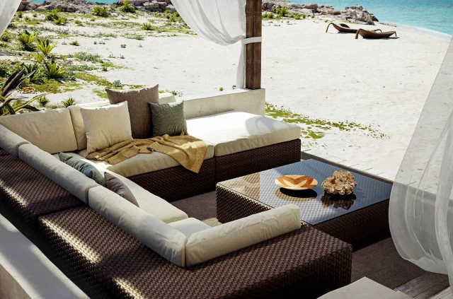 Ein moderner Outdoor-Loungebereich mit gepolsterten Korbsofas und einem gläsernen Couchtisch, der mit exquisitem Handwerk gestaltet wurde, bietet einen Blick auf einen Sandstrand inmitten von üppigem Grün. Weiße Vorhänge umrahmen den Sitzbereich und spiegeln Garten- und Landschaftsbau aus Luxemburg und Deutschland wider.