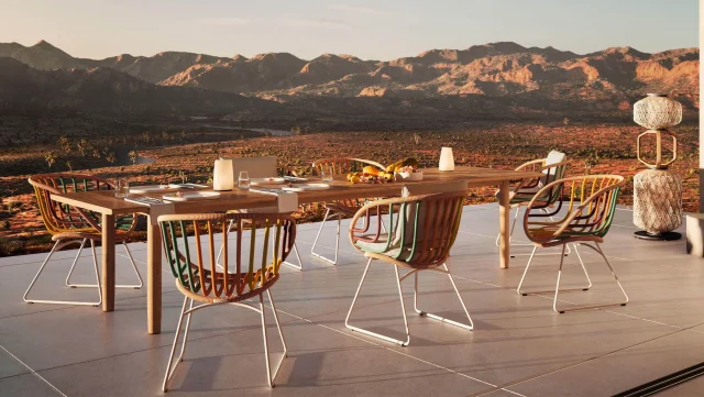 Ein moderner Outdoor-Esstisch von Burelbach mit sechs Stühlen steht auf einer gefliesten Terrasse mit Blick auf eine malerische Berglandschaft bei Sonnenuntergang. Gefertigt in handwerklicher Qualität aus Luxemburg und Deutschland, ist der Tisch für eine Mahlzeit im warmen Sonnenlicht gedeckt.