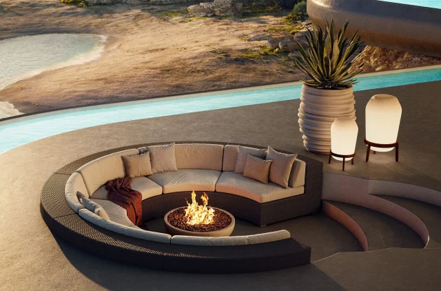 Ein moderner, versenkter Sitzbereich im Freien mit einem geschwungenen Sofa und einer zentralen Feuerstelle, umgeben von einem Pool. Stilvolle Gartenmöbel werten den Raum auf, während zwei leuchtende Lampen und eine große Topfpflanze das Gardendesign vor einer felsigen Kulisse vervollständigen.