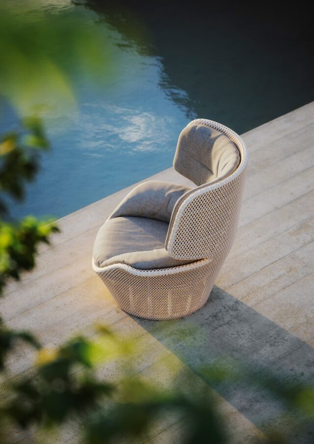 Ein gepolsterter, moderner Flechtstuhl - ein Beispiel für handwerklich gefertigte Gartenmöbel - steht auf einer Betonterrasse neben einem Gewässer, mit gedämpftem Sonnenlicht und grünem Laub im Vordergrund.
