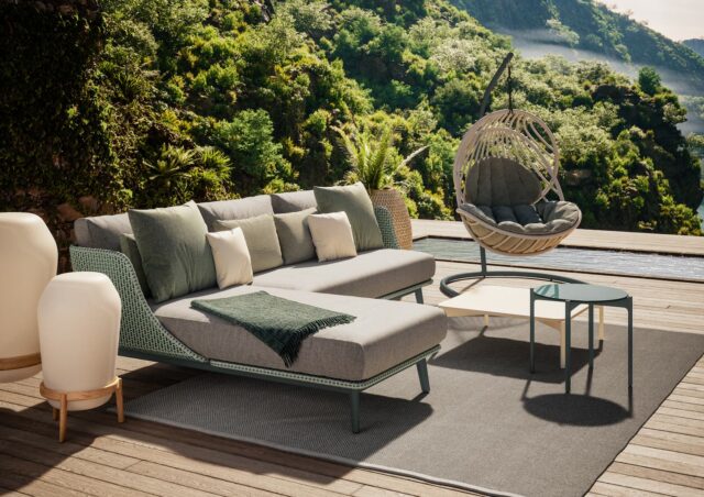 Terrasse im Freien mit einem grauen Chaiselounge-Sofa, gemütlichen Decken und Kissen, einem runden Hängesessel, kleinen Tischen und Topfpflanzen - auf einer Holzterrasse mit Blick auf üppige grüne Hügel und einen Pool. Perfekt für Gardendesign in Deutschland und Luxemburg.
