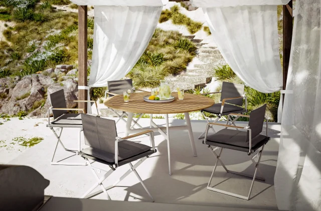 Ein runder Holztisch mit fünf modernen Stühlen steht unter einem weißen Baldachin mit transparenten Vorhängen auf einer von Garten- und Landschaftsbau Handwerk gestalteten Terrasse, umgeben von Pflanzen und Steinstufen, auf dem Tisch stehen Getränke und Zitronen.