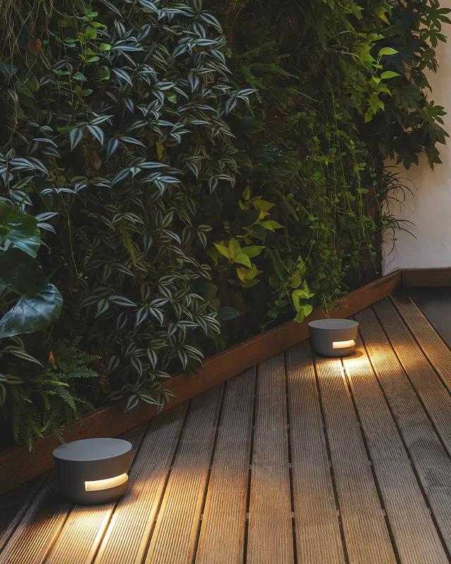Zwei runde, moderne Burelbach-Außenleuchten beleuchten eine Holzterrasse neben einem üppig grünen vertikalen Garten mit verschiedenen Blattpflanzen und werfen ein warmes Licht auf die Oberfläche - die perfekte Kulisse für stilvolle Gartenmöbel.