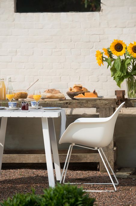 Ein weißer Tisch im Freien mit Frühstück - Saft, Marmelade und Brot - steht neben einem weißen Stuhl. Im Hintergrund stehen rustikale Gartenmöbel mit Brot, Käse und Sonnenblumen vor einer weißen Backsteinwand - eine Szene, die man in Luxemburg und Deutschland findet.