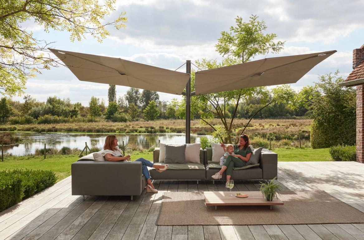 Zwei Frauen sitzen und unterhalten sich auf Sofas unter großen beigen Sonnenschirmen auf einer Holzterrasse mit Blick auf eine Graslandschaft und einen Teich - eine einladende Umgebung, die das feine Handwerk von Burelbach widerspiegelt, inspiriert von Stilen aus Luxemburg und Deutschland.