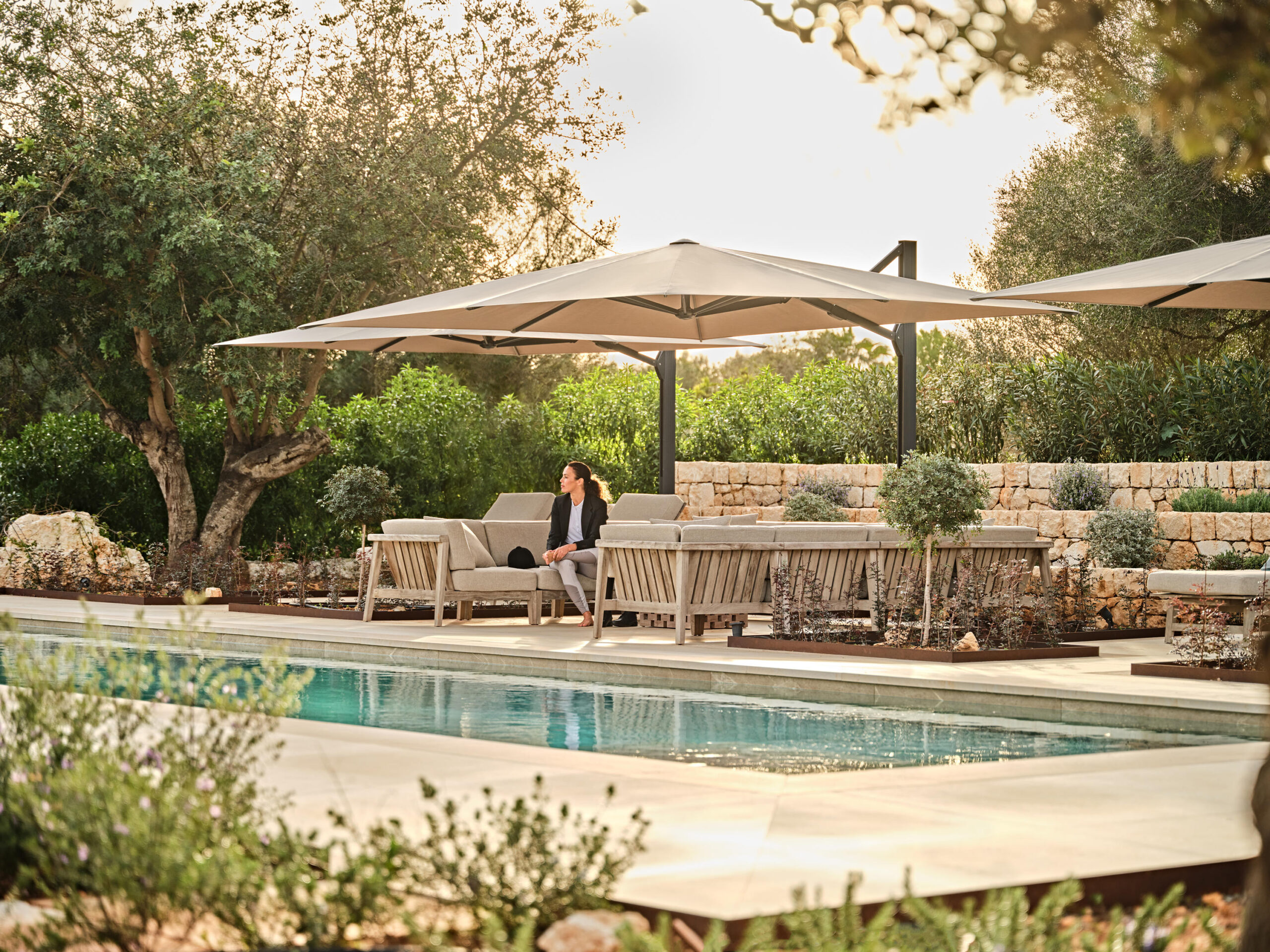 Eine Frau sitzt auf einem Liegestuhl am Pool unter einem großen Sonnenschirm, umgeben von eleganten Gartenmöbeln und Steinmauern, und genießt eine friedliche Umgebung im Freien mit üppigem Grün.