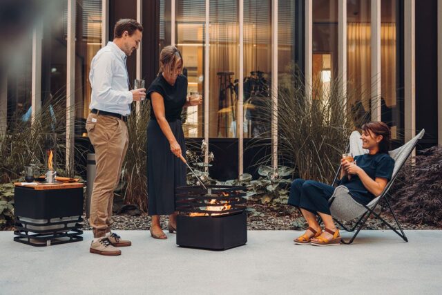 Drei Personen genießen Getränke um eine Feuerstelle auf einer Terrasse mit stilvollen Gartenmöbeln; eine Frau sitzt, während eine andere sich um das Feuer kümmert, und ein Mann steht daneben. Das hohe Gras und die modernen Burelbach-Fenster vervollständigen die Kulisse der Gartengestaltung.