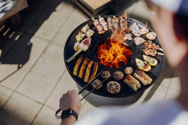 Eine Person zeigt ihr handwerkliches Geschick und grillt verschiedene Fleischsorten, Gemüse und Spieße auf einem runden Grill mit zentraler Flamme im Freien, umgeben von einer Steinterrasse in hellem Sonnenlicht - eine ideale Szene für Garten- und Landschaftsbau von Burelbach.