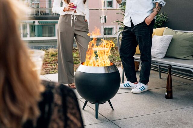 Zwei Personen stehen vor einer modernen schwarzen Feuerstelle mit hohen Flammen auf einer Dachterrasse, die stilvolle Gartenmöbel präsentiert. Eine dritte Person sitzt im Vordergrund, während im Hintergrund eine Couch mit Kissen das einladende Gartendesign ergänzt.
