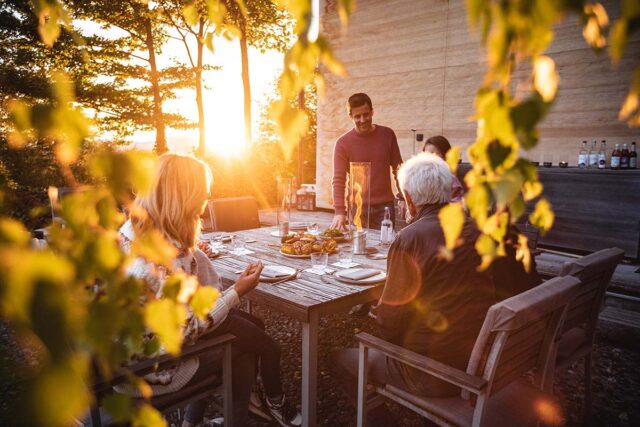 Eine Gruppe von Menschen sitzt um einen rustikalen Holztisch von Burelbach Garten- und Landschaftsbau und teilt eine Mahlzeit bei Sonnenuntergang. Goldenes Sonnenlicht fällt durch die Blätter und schafft eine warme, entspannte Atmosphäre in Deutschland und Luxemburg.