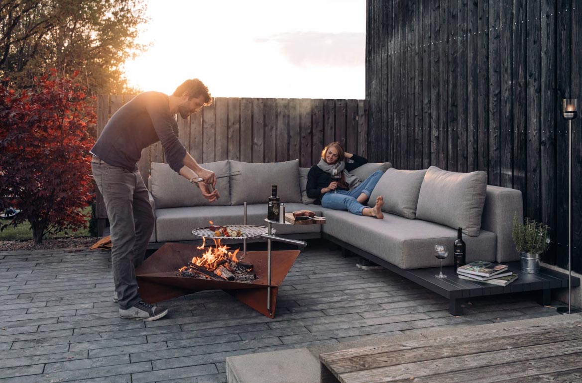 Ein Mann grillt Essen über einer modernen Feuerstelle auf einer Terrasse mit stilvollen Gartenmöbeln, während eine Frau sich auf einem grauen Outdoor-Sofa entspannt, einen Drink in der Hand hält und lächelt. Es ist Sonnenuntergang, der mit Bäumen und einem Holzzaun im Hintergrund eine gemütliche Gardendesign-Atmosphäre schafft.