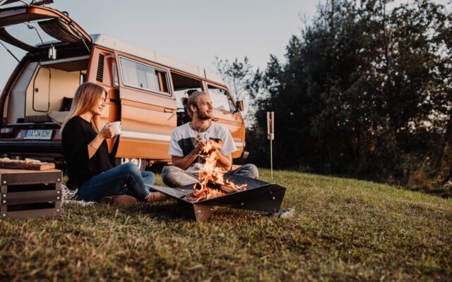 Ein Mann und eine Frau sitzen im Gras neben einem Lagerfeuer und trinken aus Bechern, während ein Lieferwagen hinter ihnen parkt. Die Szene spielt sich im Freien ab, mit Bäumen und Gartenmöbeln in der Nähe einer Kiste, die eine gemütliche Atmosphäre in der Natur schaffen.