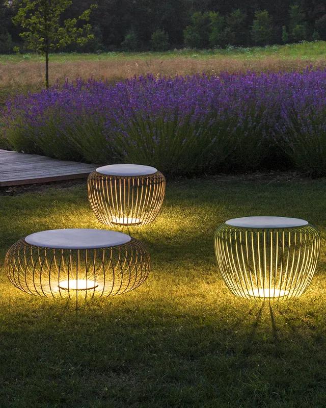 Drei moderne Außenleuchten mit Metalldrahtsockeln und flachen Aufsätzen leuchten auf einem grasbewachsenen Rasen in der Nähe eines Holzstegs und lila Blumen in der Abenddämmerung - eine einladende Szene, die Gardendesign inspiriert von Burelbach Handwerk zeigt.