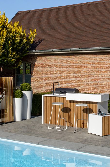 Moderne Außenküche und Barbereich mit hölzernen und weißen Schränken, Barhockern und einem Grill, neben einem Swimmingpool - ein Beispiel für Handwerk im Garten- und Landschaftsbau in Luxemburg und Deutschland - vor einem Backsteinhaus mit hohen Pflanzgefäßen und einem Baum.